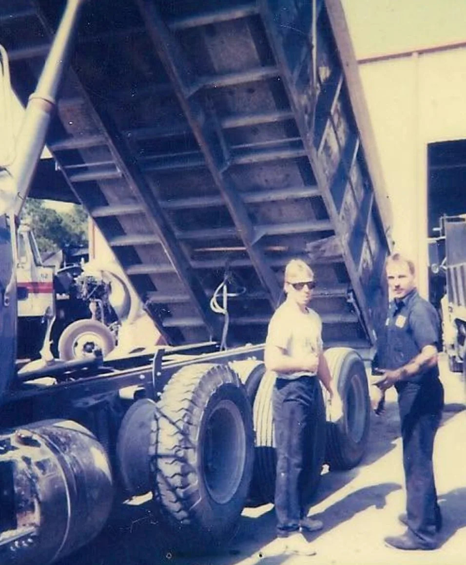 Two early PAW crew members standing beside a dump truck with its bed raised in the yard