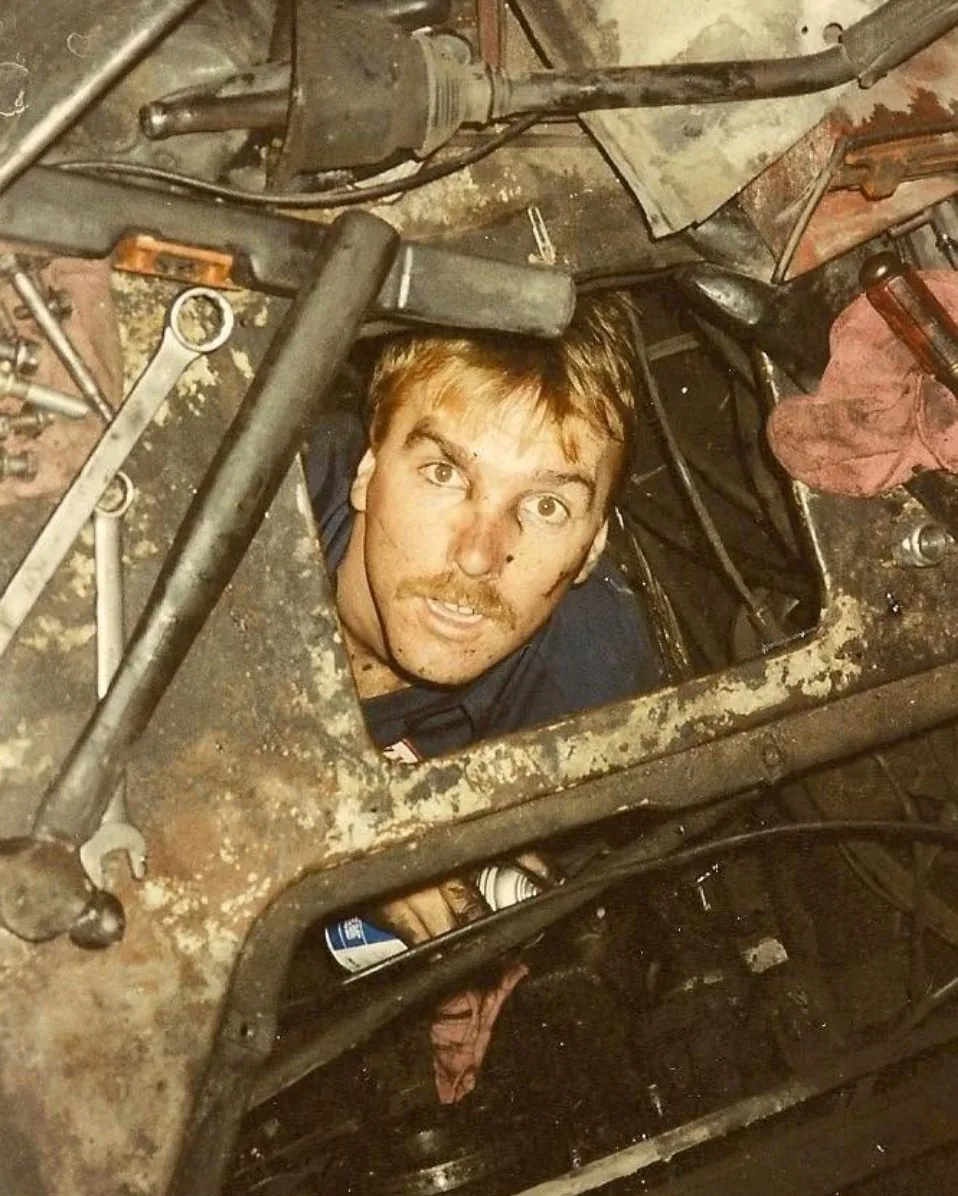 Early PAW mechanic looking up from beneath a truck chassis surrounded by tools during a repair